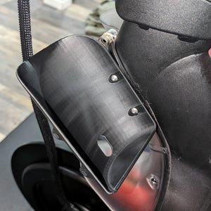 May include: A black, curved, plastic component attached to a black object, possibly a vehicle. The component has a rectangular shape with rounded edges and is secured with silver rivets. A black braided cord is visible.