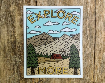May include: A sticker with a landscape design. The sticker features the words "EXPLORE MORE" in yellow, above a mountain scene with a red tent, green trees, and a blue sky with clouds. The sticker is on a wooden surface.
