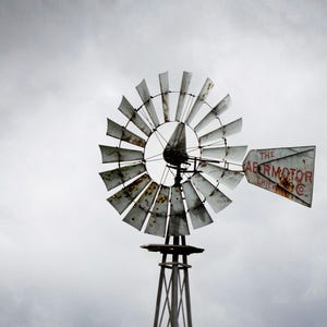 May include: A weathered windmill against a cloudy sky. The blades are made of metal and show signs of rust. The tail of the windmill has the text "THE AERMOTOR CHICAGO". The windmill is mounted on a wooden tower.
