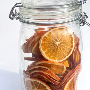 May include: A clear glass jar with a metal clasp filled with dried orange slices. The orange slices are a vibrant orange color and are arranged in a visually appealing way inside the jar. The jar is sitting on a white surface.