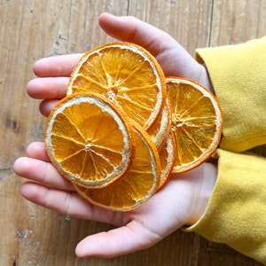 May include: A handful of dried orange slices, showcasing a vibrant orange hue with a lighter, almost white, outer ring. The slices are held against a wooden background, with a person wearing a yellow long-sleeved shirt.