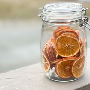 May include: A clear glass jar with a hinged lid filled with dried orange slices. The orange slices are a warm orange color and are stacked inside the jar. The jar is sitting on a light-colored wooden surface.