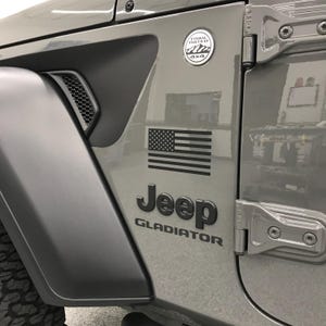 May include: Close-up of a gray Jeep Gladiator door with a black American flag decal and the Jeep logo. A Trail Rated 4x4 badge is also visible. The door has a matte finish and a black vent.