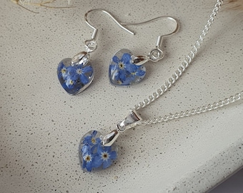 May include: A silver necklace and earrings set with heart-shaped pendants filled with pressed blue forget-me-not flowers.