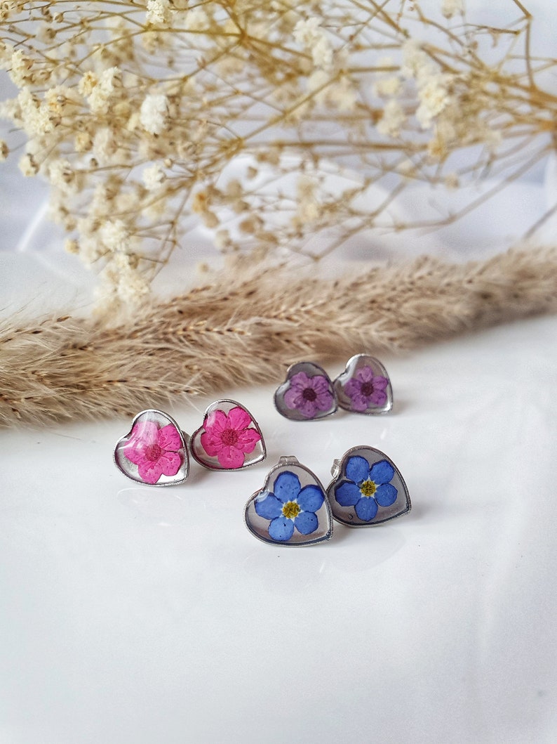 May include: Three pairs of heart-shaped earrings with pressed flowers inside. The earrings feature pink, purple, and blue flowers.