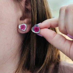 May include: A pair of heart-shaped earrings with a pink flower pressed inside each one. The earrings are silver and have a shiny finish.