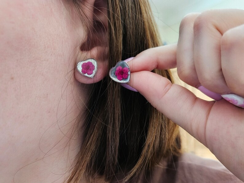 May include: A pair of heart-shaped earrings with a pink flower pressed inside each one. The earrings are silver and have a shiny finish.