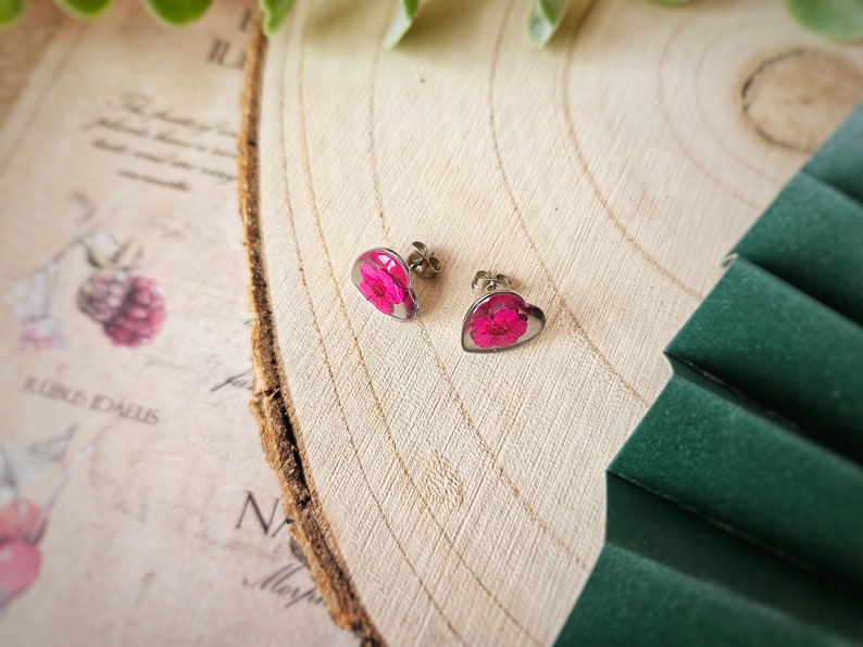 May include: A pair of heart-shaped earrings with pink pressed flowers inside. The earrings are silver and have a post back.