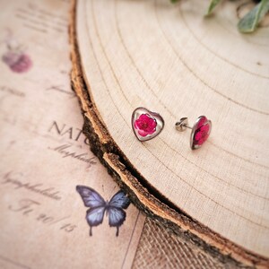May include: A pair of silver heart-shaped earrings with pink pressed flowers inside. The earrings are on a wooden surface.
