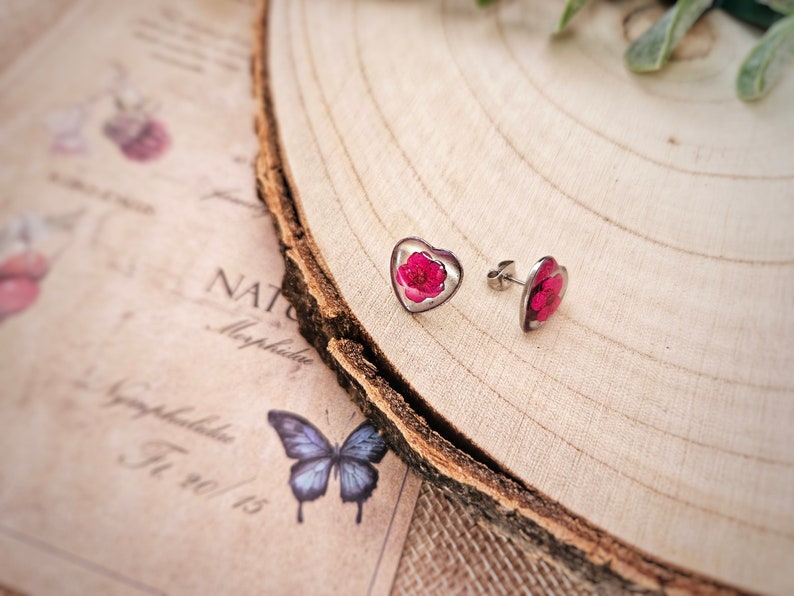 May include: A pair of silver heart-shaped earrings with pink pressed flowers inside. The earrings are on a wooden surface.