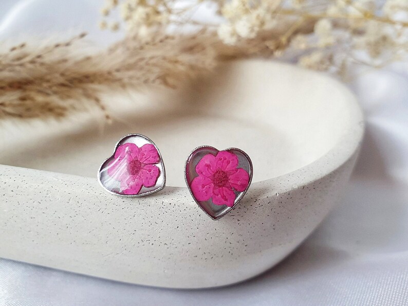 May include: A pair of heart-shaped earrings with a pink flower pressed inside each one. The earrings are silver and have a shiny finish.