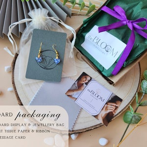 May include: A standard packaging setup for jewelry, featuring a cardboard display card with a pair of blue and gold earrings, a small white drawstring bag, a green gift box with a purple ribbon, and a business card with the text "the BLOOM jewellery".