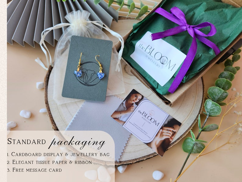 May include: A standard packaging setup for jewelry, featuring a cardboard display card with a pair of blue and gold earrings, a small white drawstring bag, a green gift box with a purple ribbon, and a business card with the text "the BLOOM jewellery".