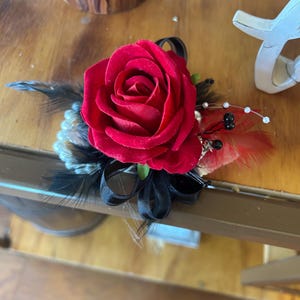 Puede incluir: Un ramillete de rosas rojas con plumas y cintas negras. El arreglo floral incluye perlas de imitaci&oacute;n y peque&ntilde;as cuentas negras. El ramillete est&aacute; dise&ntilde;ado para eventos formales y ocasiones especiales.
