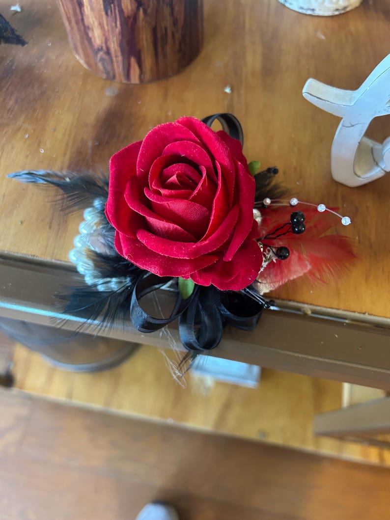 Puede incluir: Un ramillete de rosas rojas con plumas y cintas negras. El arreglo floral incluye perlas de imitaci&oacute;n y peque&ntilde;as cuentas negras. El ramillete est&aacute; dise&ntilde;ado para eventos formales y ocasiones especiales.