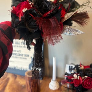 Puede incluir: Un ramo artesanal con rosas negras y rojas, acentuado con plumas negras y burdeos, y hojas plateadas con purpurina. El ramo est&aacute; adornado con broches decorativos y perlas, ideal para una boda o evento especial.
