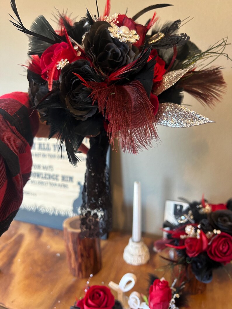 Puede incluir: Un ramo artesanal con rosas negras y rojas, acentuado con plumas negras y burdeos, y hojas plateadas con purpurina. El ramo est&aacute; adornado con broches decorativos y perlas, ideal para una boda o evento especial.