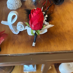 Puede incluir: Un prendedor de rosa roja con plumas negras y detalles plateados. La rosa es el punto focal, con un tallo negro envuelto en purpurina plateada. Un accesorio floral.