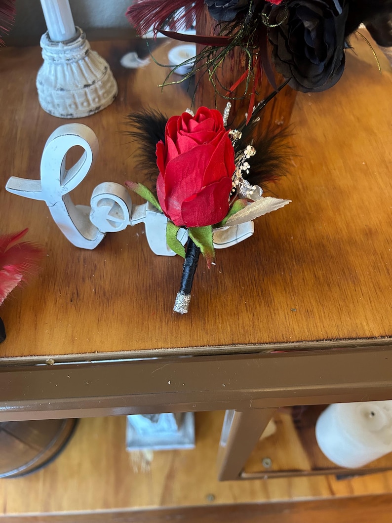 Puede incluir: Un prendedor de rosa roja con plumas negras y detalles plateados. La rosa es el punto focal, con un tallo negro envuelto en purpurina plateada. Un accesorio floral.