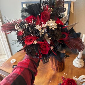 Puede incluir: Un ramo de novia con rosas rojas y negras, plumas negras y adornos decorativos. El ramo incluye una mezcla de texturas y materiales, con un prendido a juego. El arreglo est&aacute; sobre un fondo de madera.