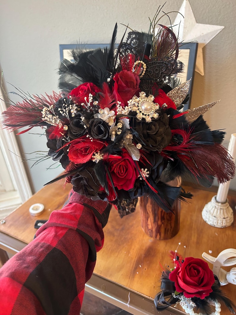 Puede incluir: Un ramo de novia con rosas rojas y negras, plumas negras y adornos decorativos. El ramo incluye una mezcla de texturas y materiales, con un prendido a juego. El arreglo est&aacute; sobre un fondo de madera.