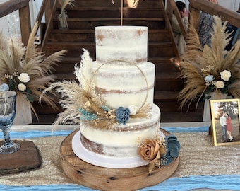 Puede incluir: Un pastel de tres niveles con glaseado blanco, decorado con hierba de la pampa seca, flores de madera azules y bronceadas, y un aro dorado. Un adorno dorado para pastel dice The Mundys. El pastel descansa sobre una base de madera.