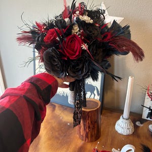 Puede incluir: Un ramo con rosas de tela negras y rosas rojas vibrantes, acentuado con plumas negras y rojas. El ramo est&aacute; adornado con detalles brillantes y una cinta de encaje negro.