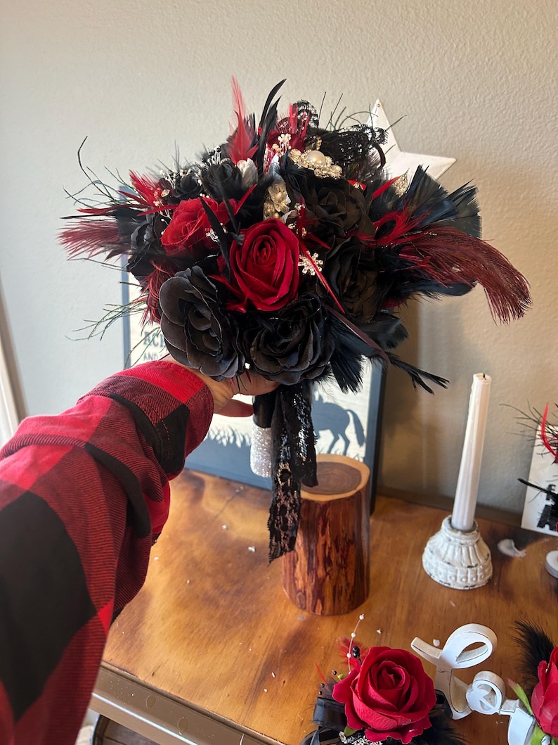 Puede incluir: Un ramo con rosas de tela negras y rosas rojas vibrantes, acentuado con plumas negras y rojas. El ramo est&aacute; adornado con detalles brillantes y una cinta de encaje negro.