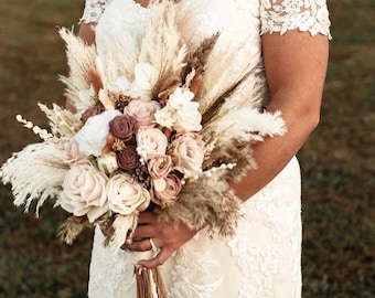 Puede incluir: Un ramo de novia con flores color crema y rosa empolvado, mezcladas con hierba de la pampa beige y follaje seco. El arreglo se sostiene frente a un vestido de encaje blanco roto con mangas cortas caídas y bordados florales.