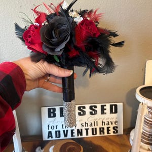 Puede incluir: Un ramo artesanal con rosas rojas y negras, acentuado con plumas negras y adornos brillantes. El mango est&aacute; envuelto en tela negra con una base de purpurina plateada. Una pieza decorativa para bodas o eventos especiales.