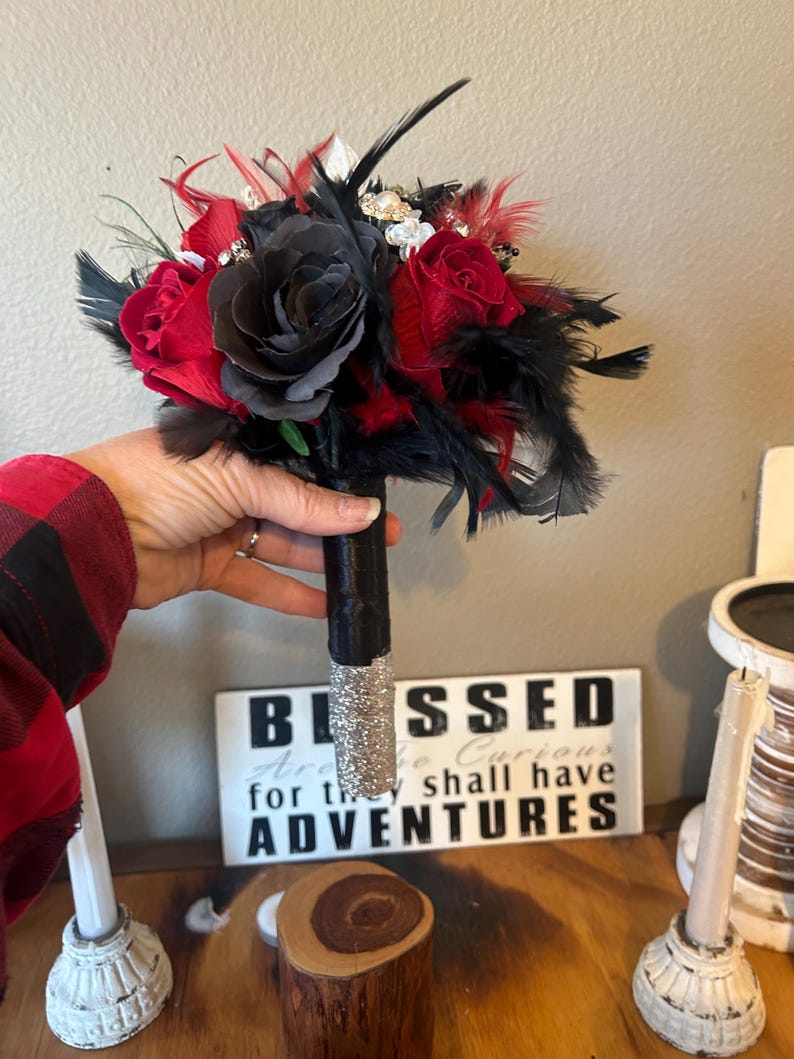 Puede incluir: Un ramo artesanal con rosas rojas y negras, acentuado con plumas negras y adornos brillantes. El mango est&aacute; envuelto en tela negra con una base de purpurina plateada. Una pieza decorativa para bodas o eventos especiales.