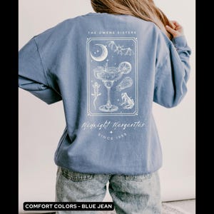 May include: A blue jean sweatshirt with a white graphic design of a margarita glass with a crescent moon, bats, and frogs. The text "The Owens Sisters Midnight Margaritas Since 1998" is printed on the sweatshirt.
