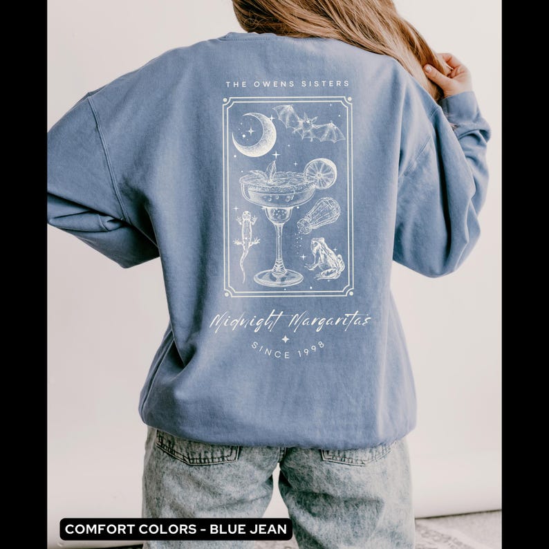 May include: A blue jean sweatshirt with a white graphic design of a margarita glass with a crescent moon, bats, and frogs. The text "The Owens Sisters Midnight Margaritas Since 1998" is printed on the sweatshirt.