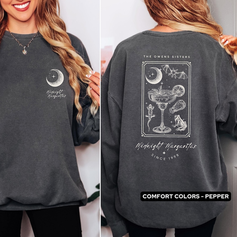 May include: A dark gray sweatshirt with a white graphic design on the front and back. The graphic features a crescent moon, stars, a bat, a frog, and a margarita glass. The text on the sweatshirt reads "Midnight Margaritas" and "The Owens Sisters Since 1998". The sweatshirt is labeled "Comfort Colors - Pepper".
