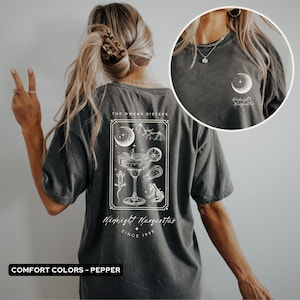 May include: Dark grey t-shirt with a white graphic on the back featuring a margarita glass, moon, bats, and the text "Midnight Margaritas Since 1998." The shirt also has a small moon and text on the front. The shirt is a comfort color.