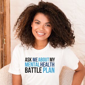 May include: White t-shirt with the text "Ask me about my mental health battle plan" in blue and black lettering.