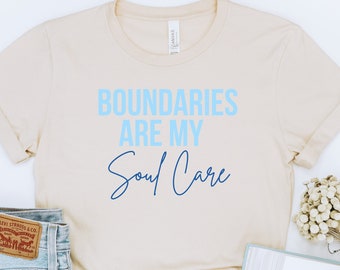 May include: A beige t-shirt with the text "Boundaries are my Soul Care" in blue and light blue lettering.