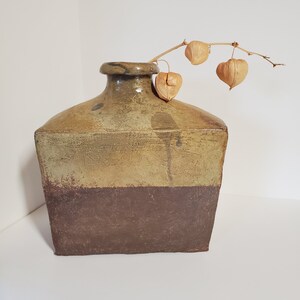 May include: A square, brown and green ceramic vase with a textured surface. The vase is filled with a single stem of dried flowers.