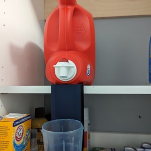 Può includere: Una bottiglia di detersivo in plastica rossa con un beccuccio bianco è montata su uno scaffale in plastica blu con un bicchiere di plastica trasparente sotto. Lo scaffale è attaccato a un armadio bianco.