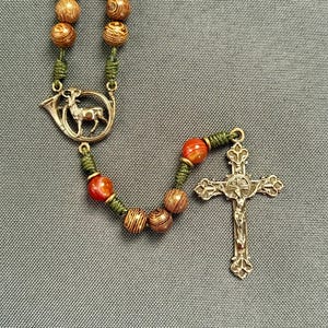 May include: A rosary with wooden beads and a silver-toned crucifix. The beads are a mix of brown and orange, strung on green cord. A silver-toned charm with a deer inside a horn is also present.