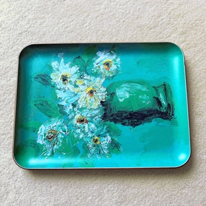 May include: A teal blue tray with a painted design of white flowers in a green vase. The tray has a slightly raised rim.