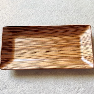 Puede incluir: Una bandeja rectangular de madera con un patrón de rayas de color marrón claro y marrón oscuro. La bandeja tiene un borde ligeramente elevado.