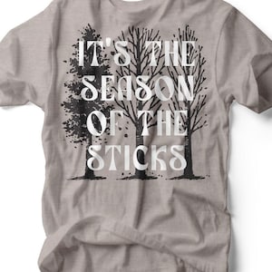 Pode incluir: Camiseta cinza mesclada com o texto branco "IT'S THE SEASON OF THE STICKS" e um gráfico de árvores pretas. A camiseta tem gola redonda e mangas curtas. A marca "RUBY'S RUBBISH" está impressa na parte inferior.