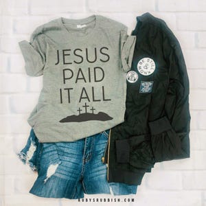 Puede incluir: Un conjunto casual que incluye una camiseta gris con la frase "JESUS PAID IT ALL" y tres cruces, combinada con shorts de mezclilla desgastados y una chaqueta bomber negra con parches. Sobre un fondo de ladrillos blancos.