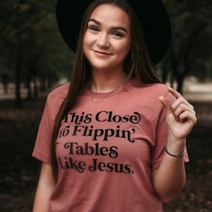 Pode incluir: Camiseta rosa empoeirado com o texto preto "This Close to Flippin' Tables Like Jesus". A pessoa na imagem está usando um chapéu preto e uma pulseira de prata.