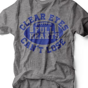 May include: A heather gray t-shirt with a blue football graphic. The graphic includes the text "CLEAR EYES, FULL HEARTS, CAN'T LOSE" in a circular arrangement around the football shape. The shirt is made of a soft material.
