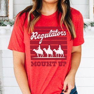 May include: Red t-shirt featuring the text "Regulators MOUNT UP" in white script, with a graphic of three camels. The top has a crew neck and short sleeves.