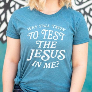 Könnte beinhalten: Türkisfarbenes T-Shirt mit dem weißen Text "WHY Y'ALL TRYIN' TO TEST THE JESUS IN ME?". Das Shirt ist an der Taille geknotet und wird mit einer Jeans getragen. Das Bild ist von der Brust aufwärts aufgenommen.