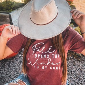 May include: A light grey fedora hat with a brown band is held up. A maroon t-shirt with the text "Fall Opens The Window To My Soul" is visible. The person is wearing blue denim shorts.