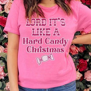 May include: Pink t-shirt with the phrase "LORD IT'S LIKE A Hard Candy Christmas" in a decorative font. A candy graphic is below the text. The shirt is worn with blue jeans.